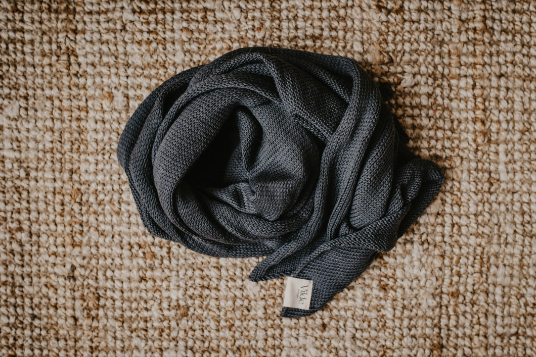Auf dem Bild ist ein Schal in der Farbe anthrazit zu sehen. Der Schal liegt auf einem beigen Hintergrund. Unten rechts am Schal sieht man das Vilde Joranger Logo. Der Schal ist glatt gestrickt, schlicht und zeitlos. Der Schal ist Handgemacht und hat einen schönen dunkel graue Farbe. 