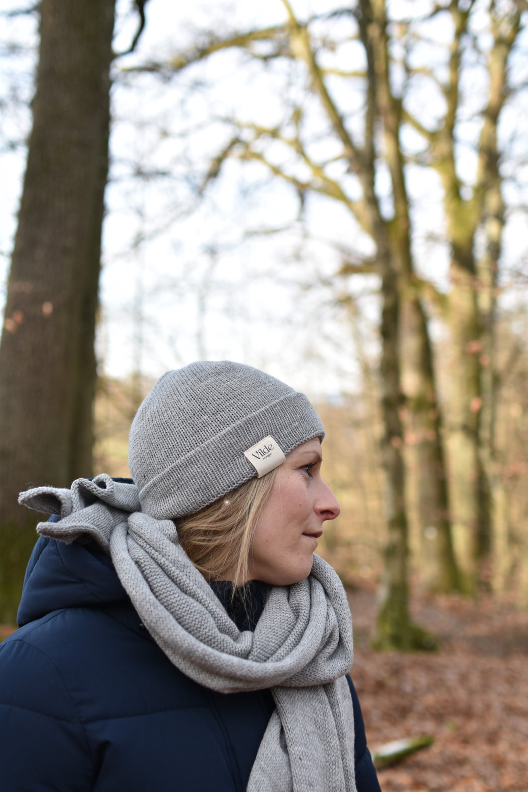 Eine Frau trägt die Natur-graue Mütze im Wald. Sie trägt auch den Schal in der Farbe Natur grau. Sie schau nach links in dem Wald rein. Auf der rechte Seite sieht man das Vilde Joranger Logo.